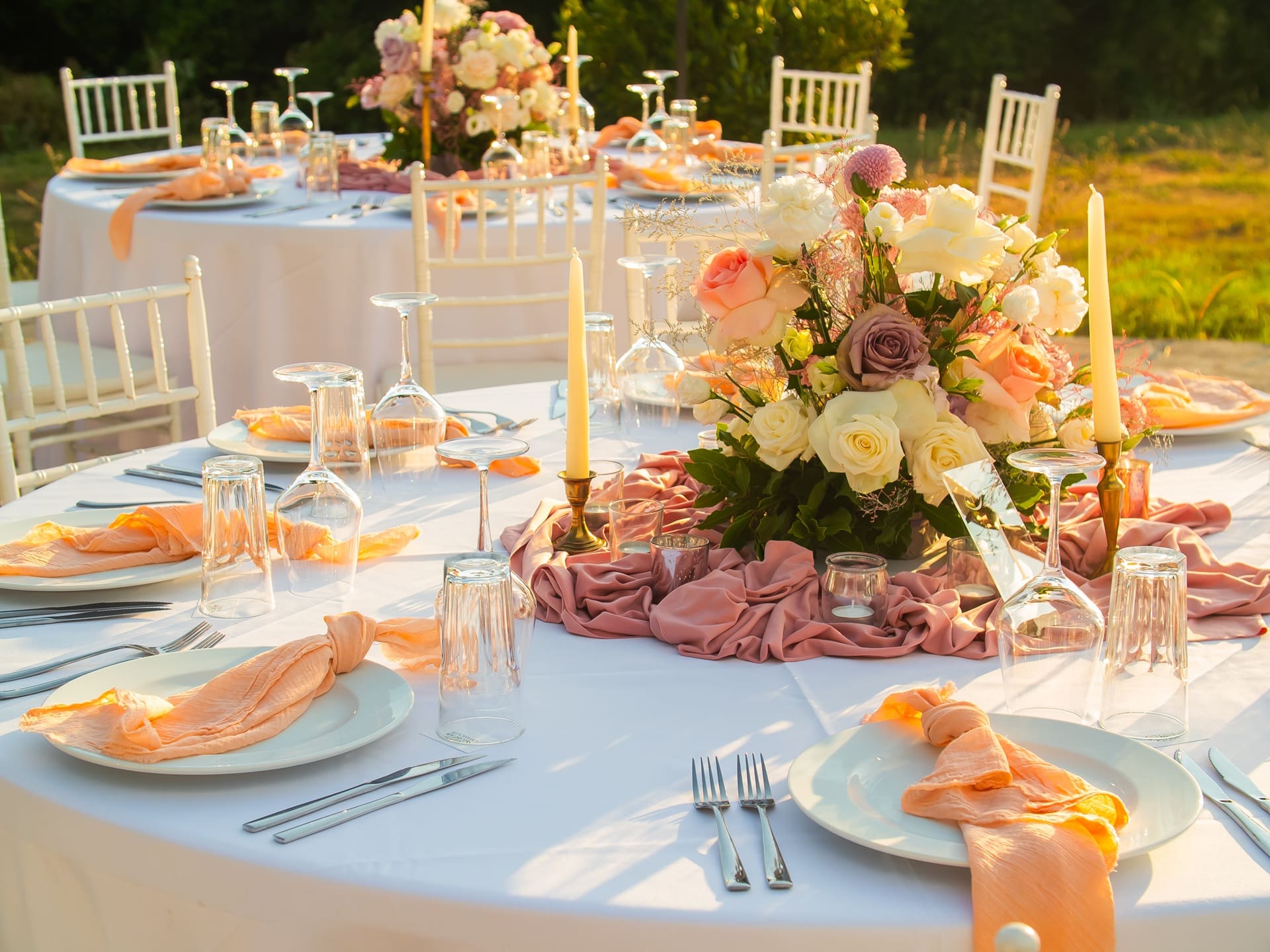 A round table set for an outdoor event with white chairs, pink and white floral centerpiece, candles, glassware, and peach napkins arranged on white tablecloths.