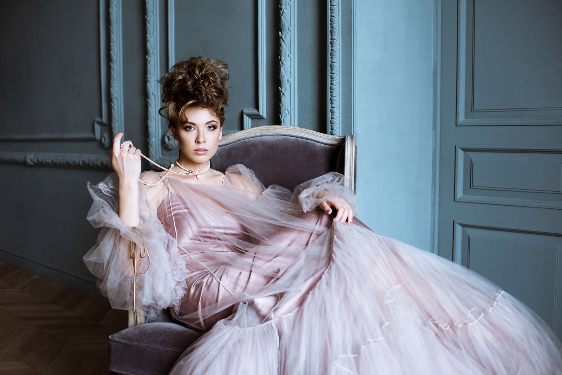 A woman in a voluminous, light pink gown sits in an ornate gray chair, holding a pearl necklace, against an elegant blue-toned wall.