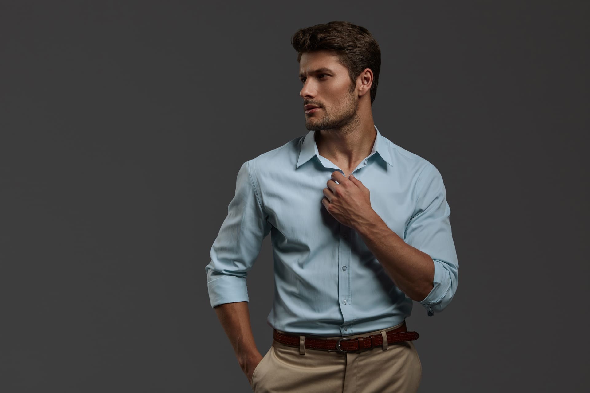 A man in a light blue dress shirt and beige pants stands against a plain dark background, looking to the side with one hand adjusting his collar.