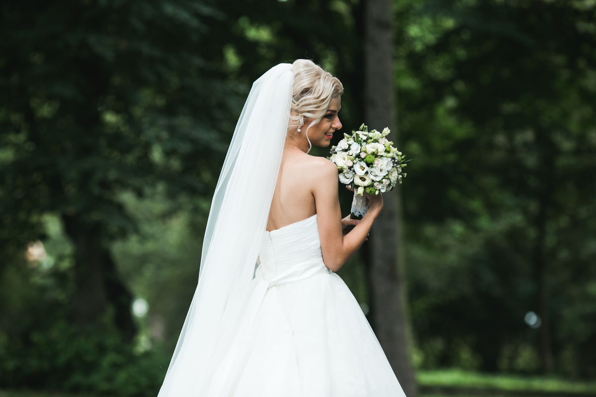 Bride in a white strapless gown and veil holding a bouquet of white flowers, standing outdoors in a green, wooded area.