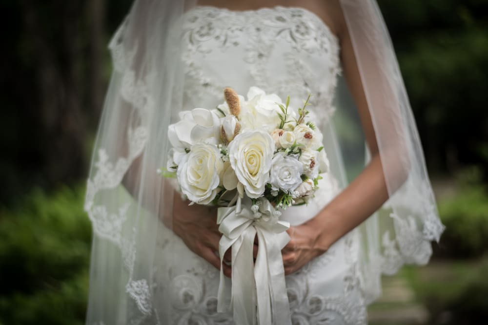 A bride in a white lace dress holds a bouquet of white flowers and greenery, standing outdoors with a veil draped over her shoulders.