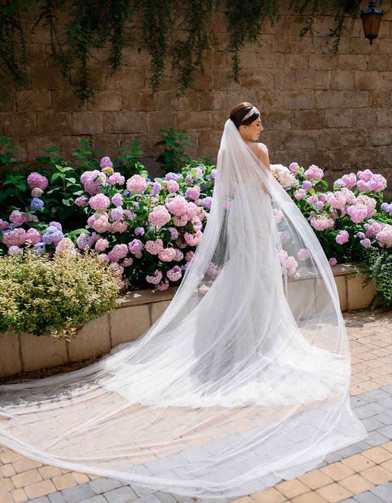 A bride in a white dress with a long veil stands on a paved path in front of blooming pink hydrangeas and a stone wall.