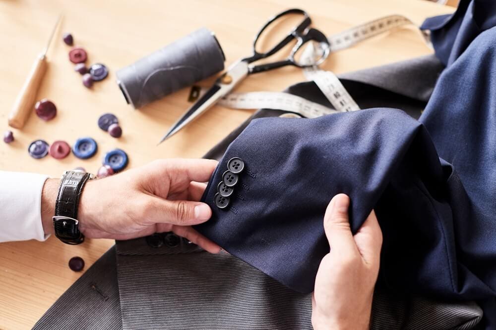 A tailor sews buttons onto a navy suit jacket, with scissors, thread, tape measure, and assorted buttons on a wooden table.