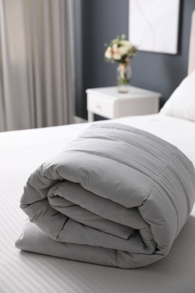A neatly folded gray comforter sits on a made bed in a modern bedroom, with a nightstand and vase of flowers in the background.