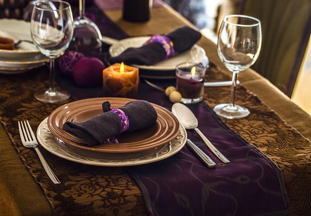 Elegant table setting with brown and purple plates, wine glasses, cutlery, candles, and dark napkins wrapped with purple holders on a decorative tablecloth.