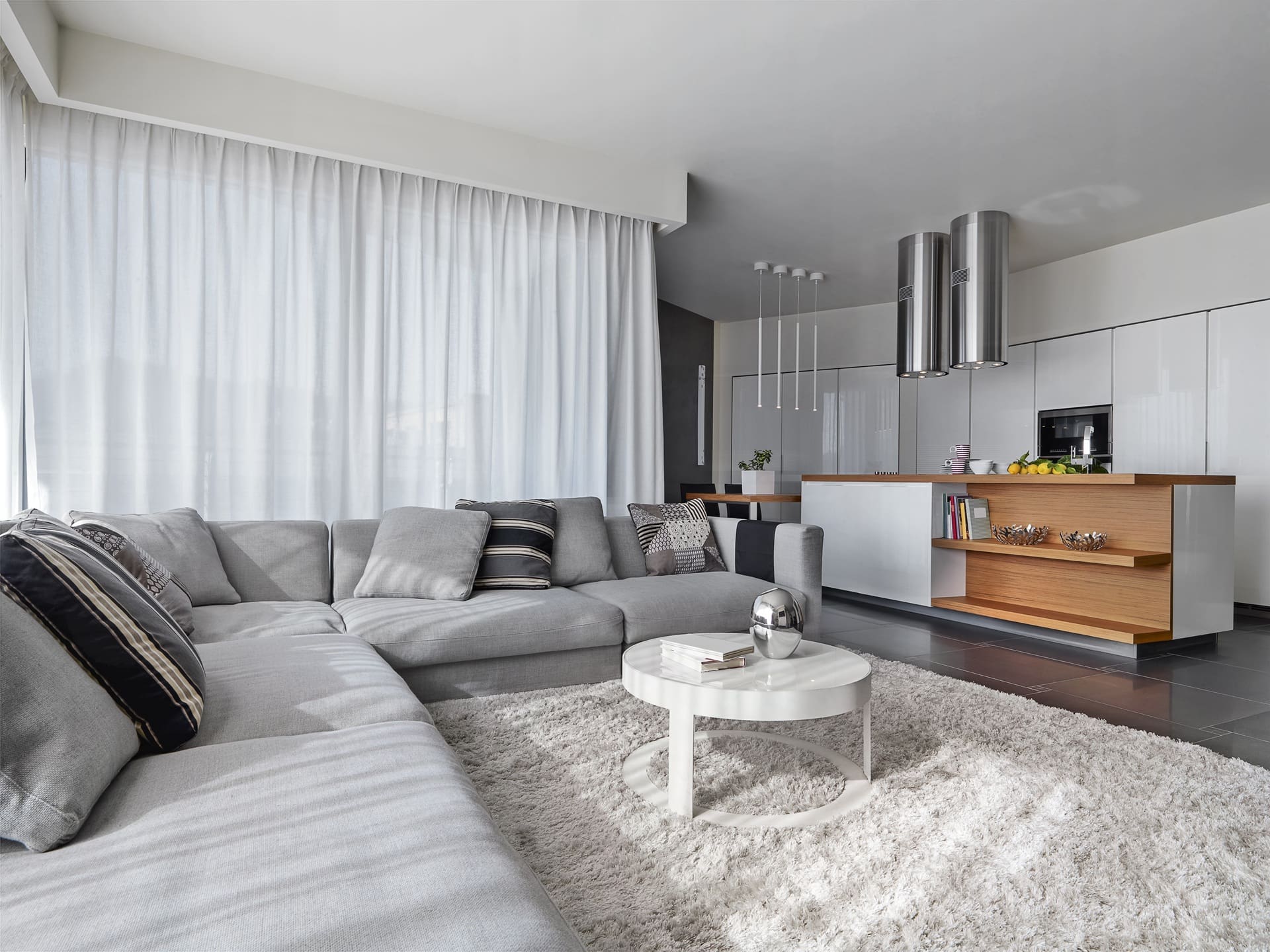 Modern open-plan living room and kitchen with a grey sectional sofa, round white coffee table, shaggy rug, and white cabinetry; natural light filters through sheer curtains.