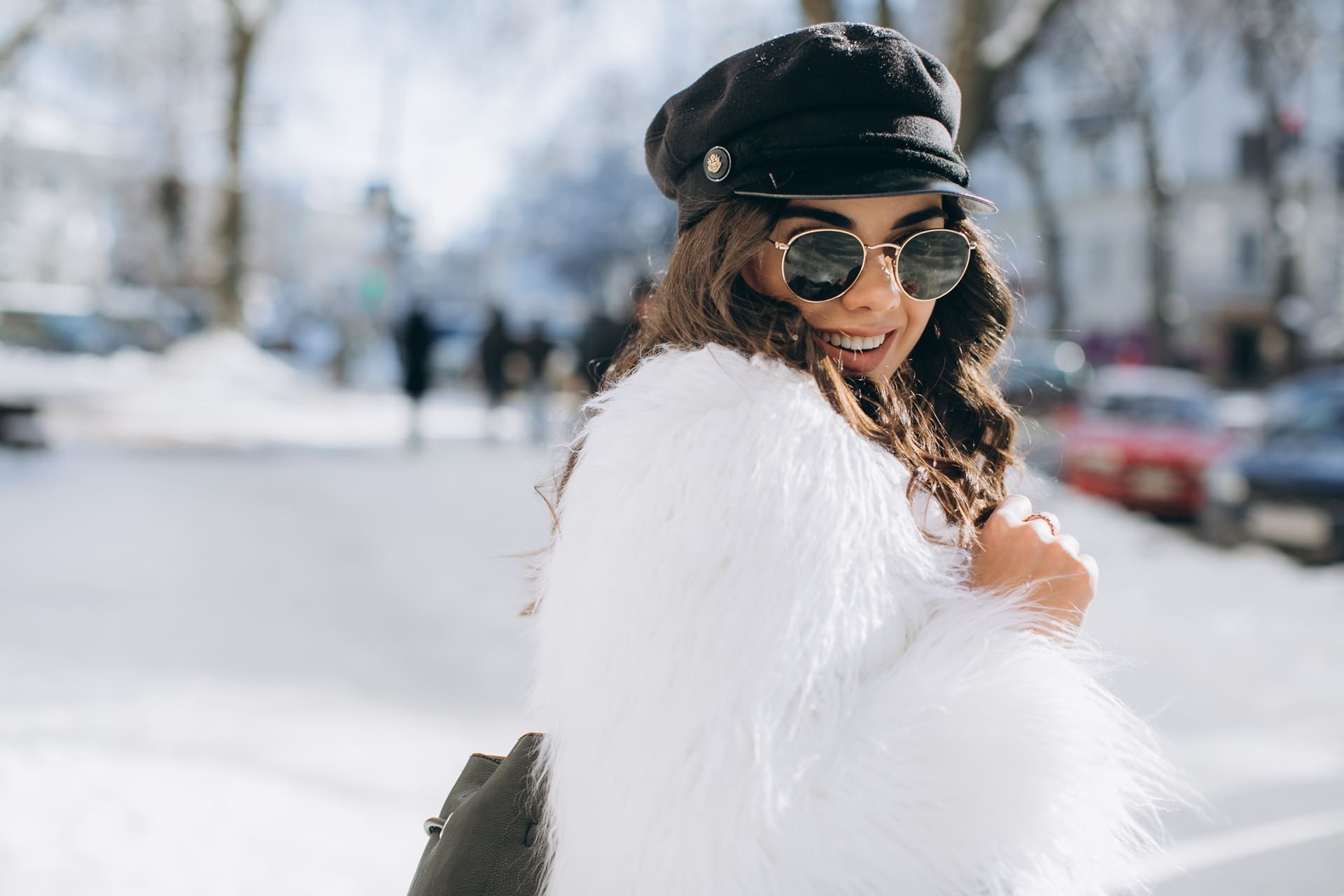 Woman wearing sunglasses, a black cap, and a white faux fur coat stands outdoors on a snowy city street, looking back and smiling.