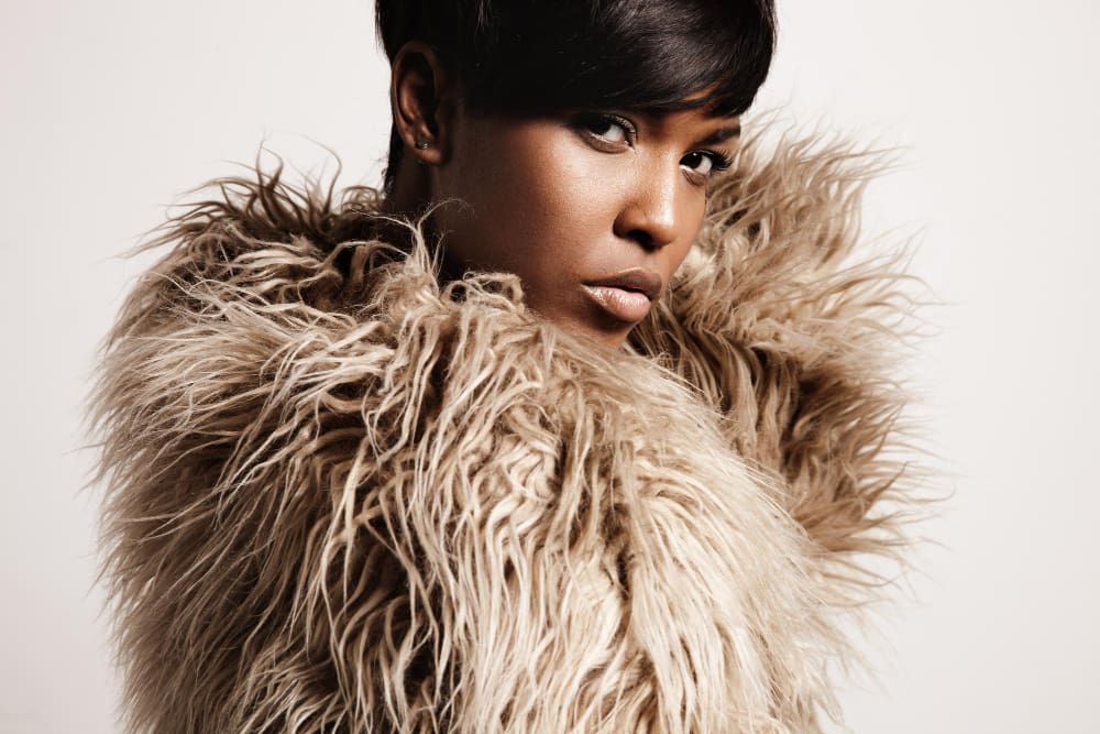 Person with short dark hair wearing a large, light brown faux fur coat looks over their shoulder against a plain background.