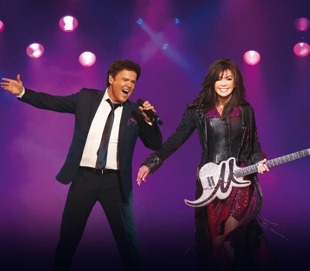 A man in a suit sings into a microphone while a woman in a dark dress holds a decorative guitar on a purple-lit stage.