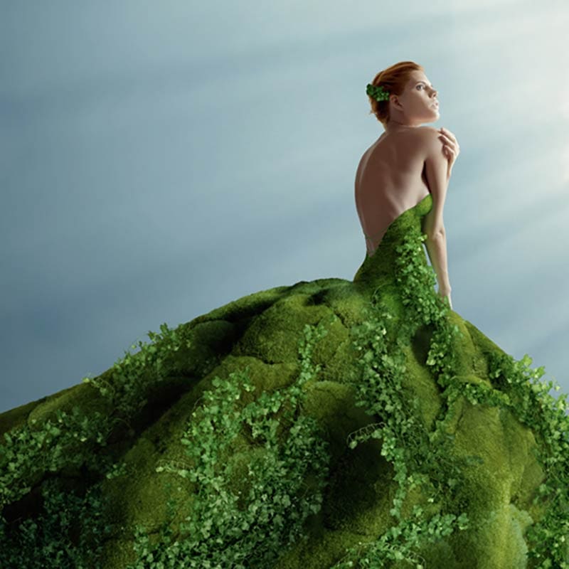A woman with red hair sits in profile, wearing a strapless green dress that appears to be made of moss and leaves, against a softly lit, blue-gray background.