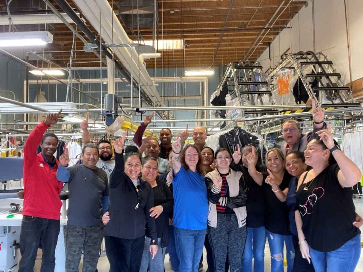 A group of people stands together inside a laundromat or dry cleaning facility, smiling and raising one finger, with clothing racks and machines visible in the background.