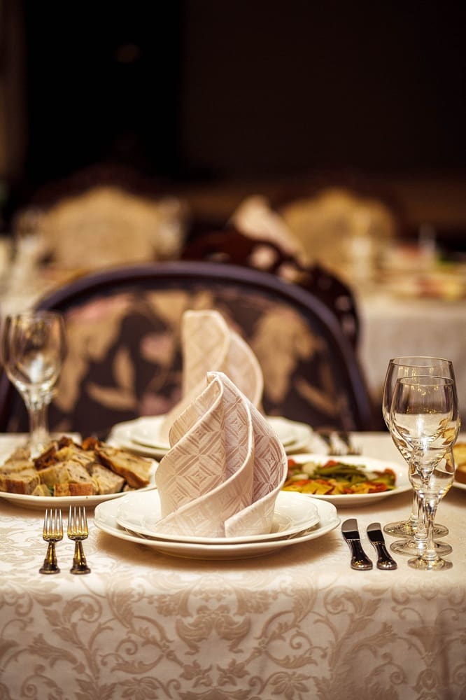 A formally set dining table with a folded napkin on a plate, bread, salad, wine glasses, and cutlery on a cream tablecloth.