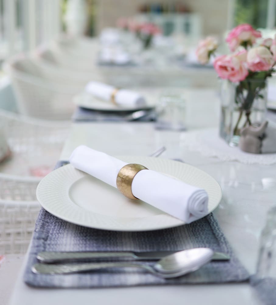 A neatly set dining table with a white plate, rolled napkin in a gold ring, silver cutlery, a glass, and a vase of pink flowers.