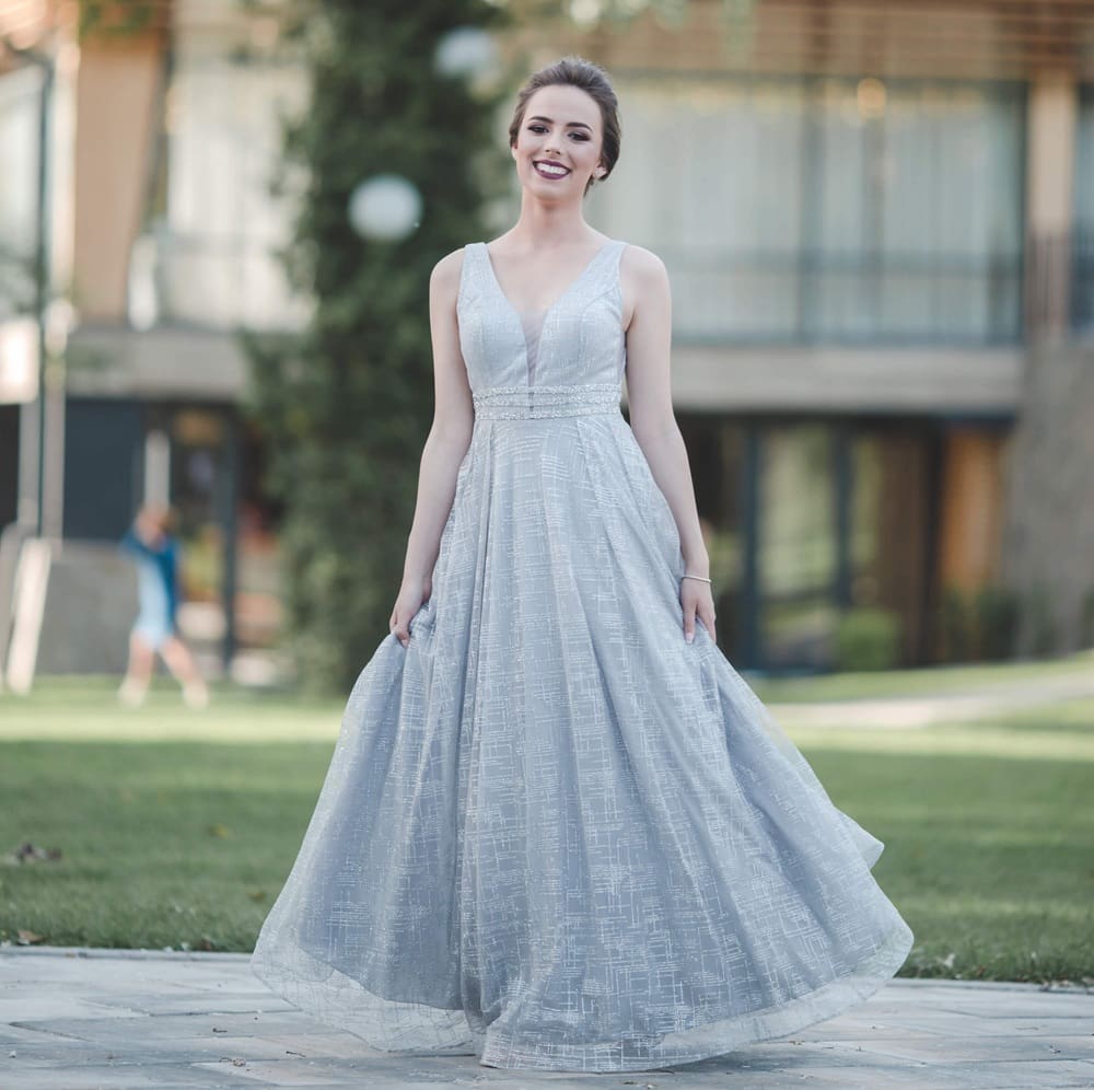 A woman stands outdoors in a sleeveless, floor-length silver gown, smiling and holding the sides of her dress. A blurred building and greenery are in the background.