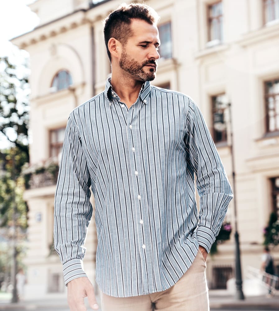 Man wearing a blue and white striped button-up shirt stands outdoors in front of a light-colored building on a sunny day.