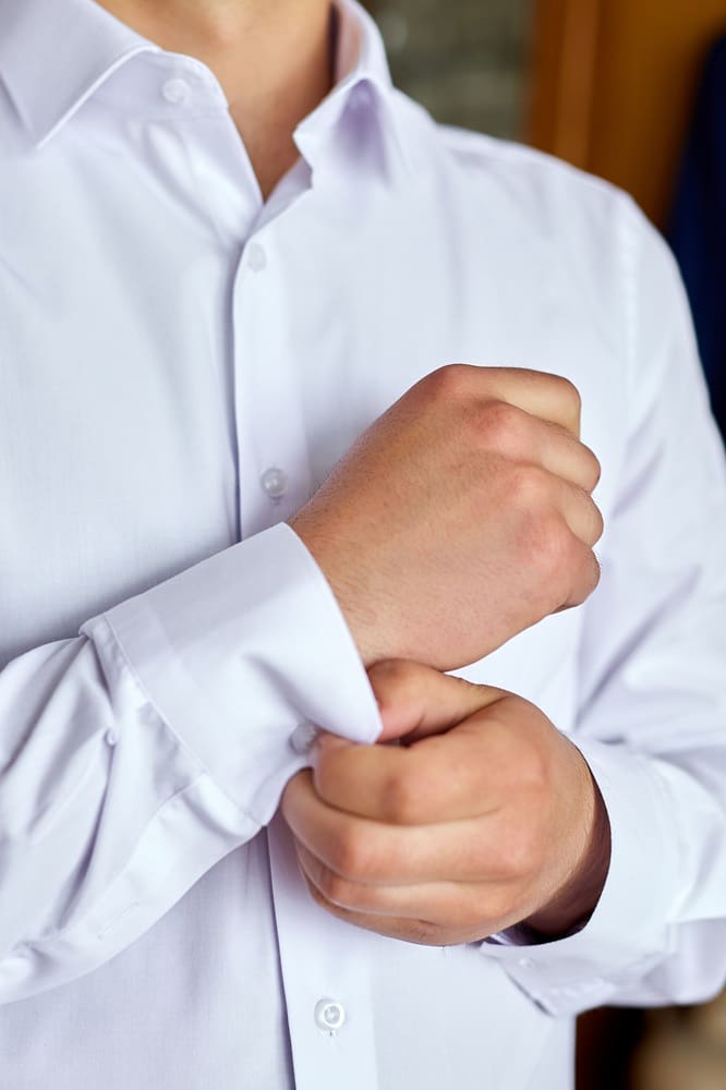 A person fastens the cuff of a white dress shirt with one hand while wearing it.