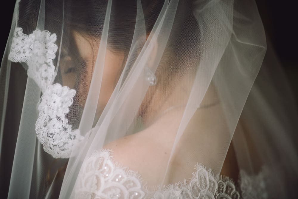 A woman in a white lace wedding dress is partially covered by a sheer veil, with lace details visible. Her face is turned slightly away from the camera.