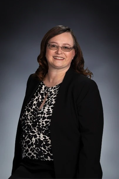A woman with shoulder-length brown hair, glasses, and a black blazer over a patterned blouse, smiling against a plain dark background.