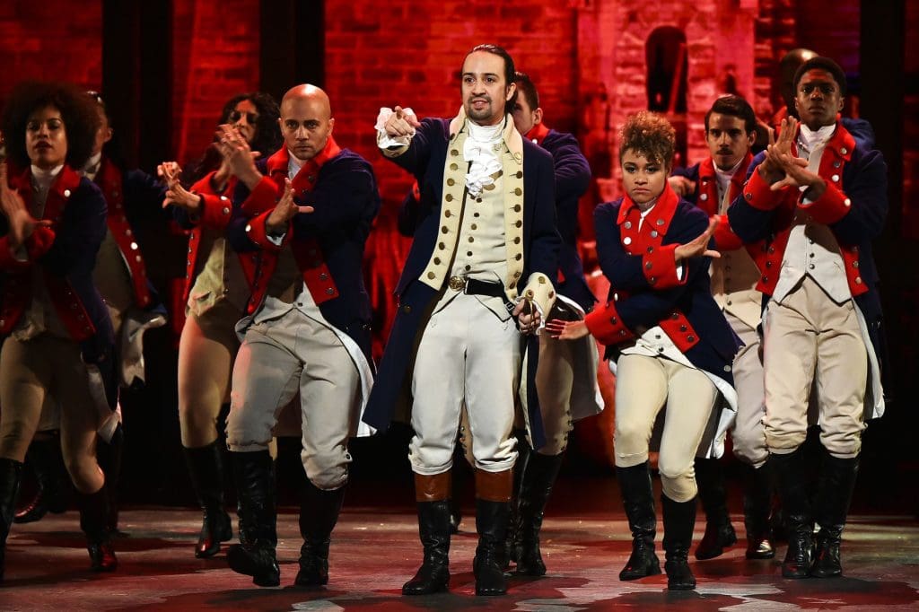 A group of performers in 18th-century military costumes dance energetically on stage with a brick wall backdrop.