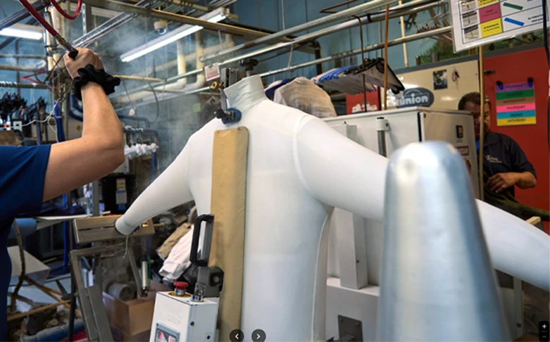 A headless mannequin dressed in a white bodysuit is being steamed in a laundry or garment care facility, with machines and workers in the background.