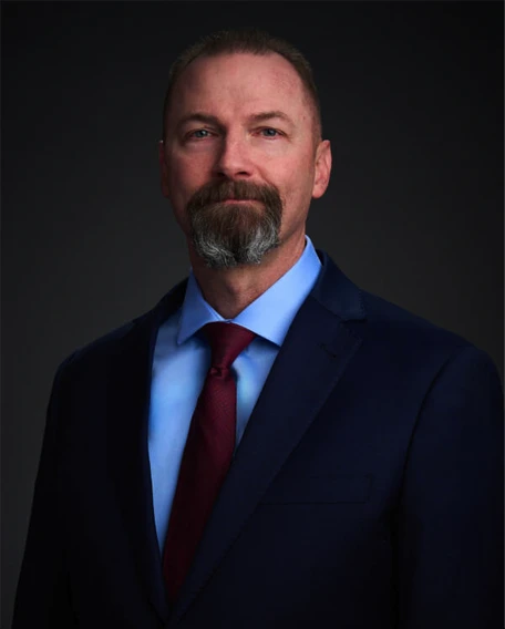 A man with short hair and a gray goatee is wearing a navy suit, light blue shirt, and burgundy tie, posing against a dark gradient background.