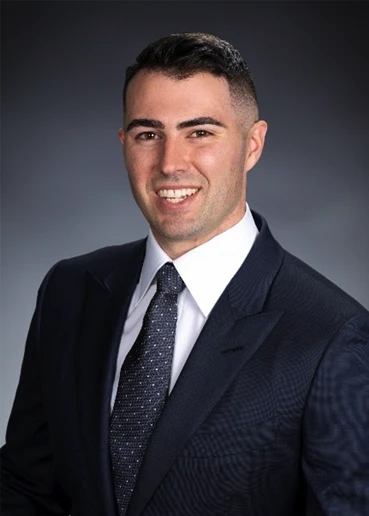 A man in a dark suit, white shirt, and patterned tie poses for a professional studio portrait against a plain gray background.