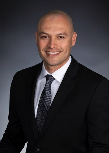 A man in a black suit, white dress shirt, and patterned tie is smiling at the camera against a plain dark background.