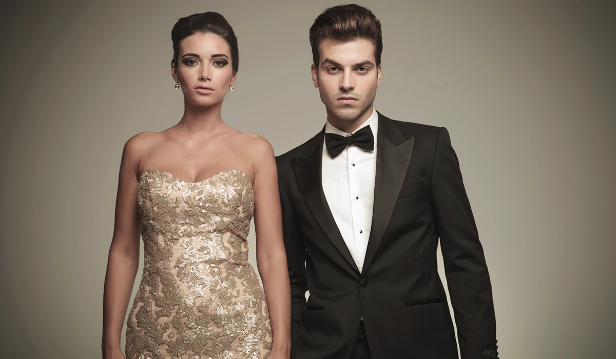 A woman in a strapless gold patterned dress stands next to a man in a black tuxedo with a bow tie, both facing forward against a plain background.