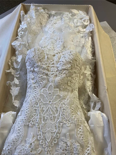 A white lace wedding dress with intricate floral and geometric patterns is displayed in a box with tissue paper.