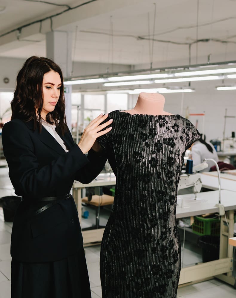 A woman adjusts a sequined black dress on a mannequin in a garment workshop with sewing machines in the background.