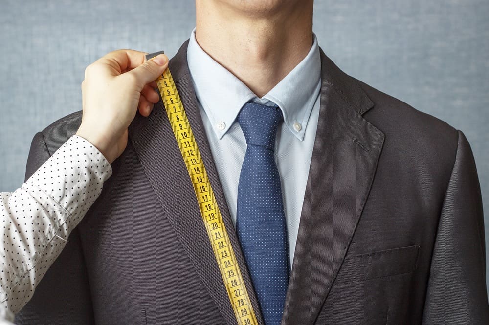 A person in a suit and tie is being measured along the shoulder with a yellow measuring tape by another person's hand.