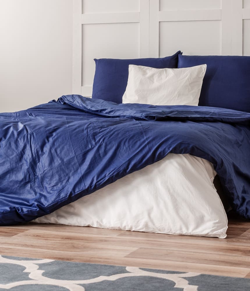 A neatly made bed with a blue duvet, blue pillows, and a white pillow, set against a white paneled wall on a wooden floor.
