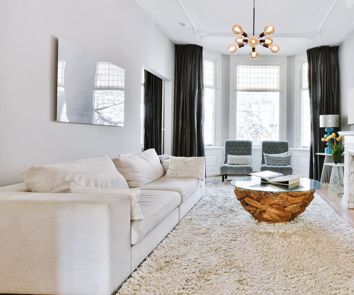 Modern living room with a large white sofa, textured rug, unique wooden coffee table, two armchairs by tall windows, and a chandelier.