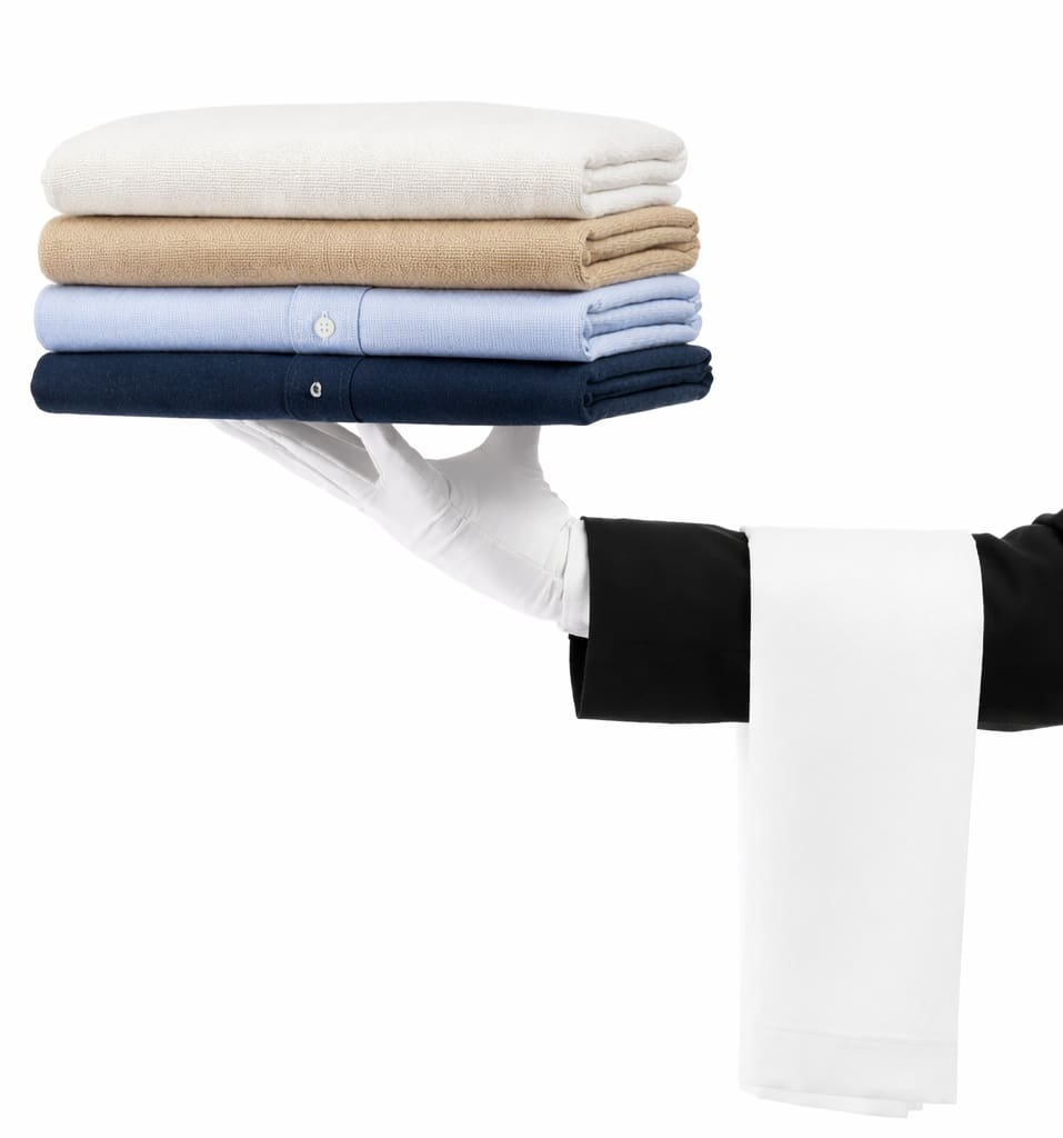 A gloved hand holds a neatly folded stack of four shirts in white, beige, light blue, and dark blue, with a white cloth draped over the arm.