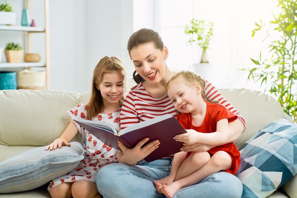 An adult and two young children sit on a sofa, smiling and reading a book together in a brightly lit living room.