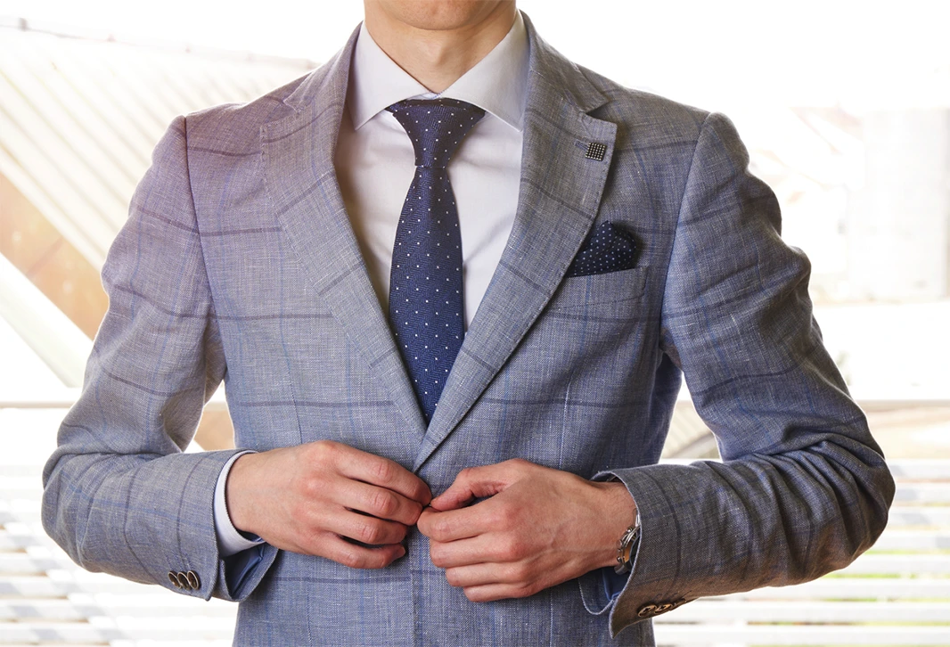 A man in a light gray suit with a white shirt and navy blue polka dot tie buttons his jacket.
