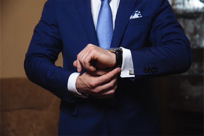 A person in a blue suit and tie adjusts a wristwatch on their left wrist, with only the upper body and arms visible.