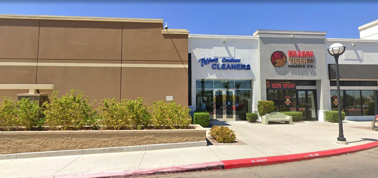 A shopping plaza with a dry cleaners called Nelson Custom Cleaners and a noodle restaurant called Dragon Tiger Noodle Co.; both storefronts face a sidewalk and parking curb.