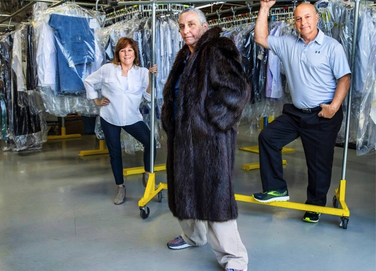 Three adults pose in a dry cleaning facility; one wears a fur coat, while the other two stand by clothing racks with laundry hanging in plastic covers.
