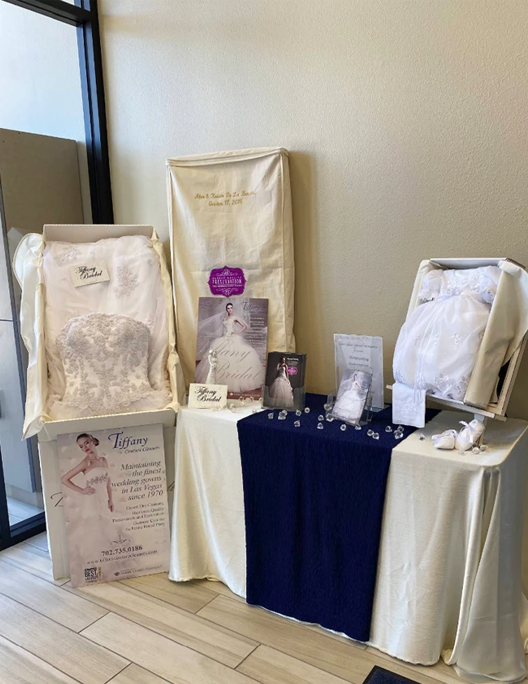 A display of preserved wedding dresses in clear boxes with informational signs and memorabilia, set on tables covered with cream and navy blue cloths near a window.