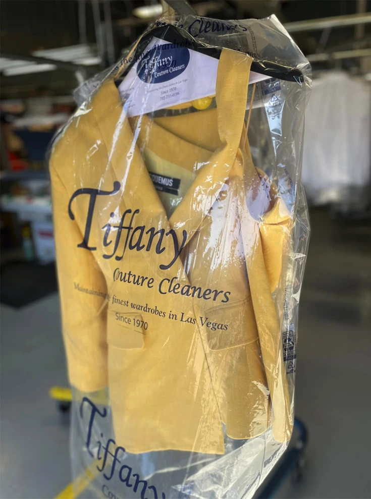 A yellow suit is hanging on a rack, covered in a transparent garment bag labeled "Tiffany Couture Cleaners" inside a dry cleaning facility.