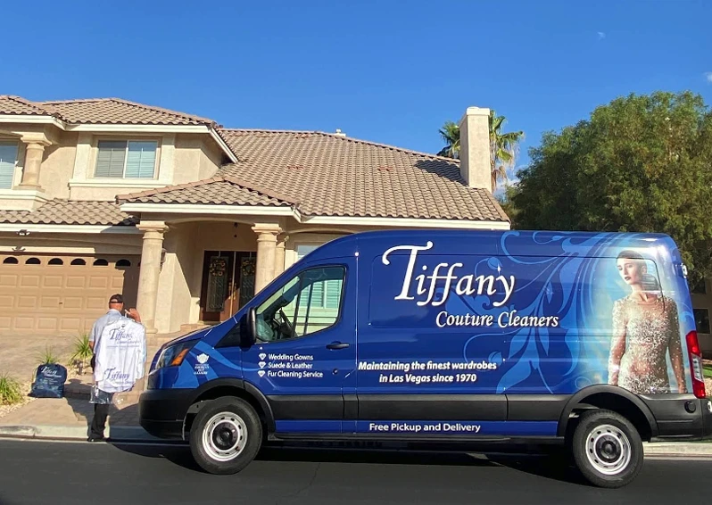 A Tiffany Couture Cleaners van is parked in front of a house, with an employee carrying garments towards the home on a sunny day.