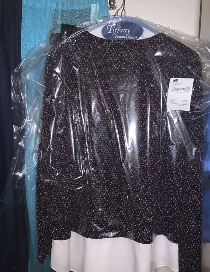 A black and white patterned blouse covered in a plastic dry-cleaning bag hangs on a hanger labeled "Tiffany Custom Cleaners.