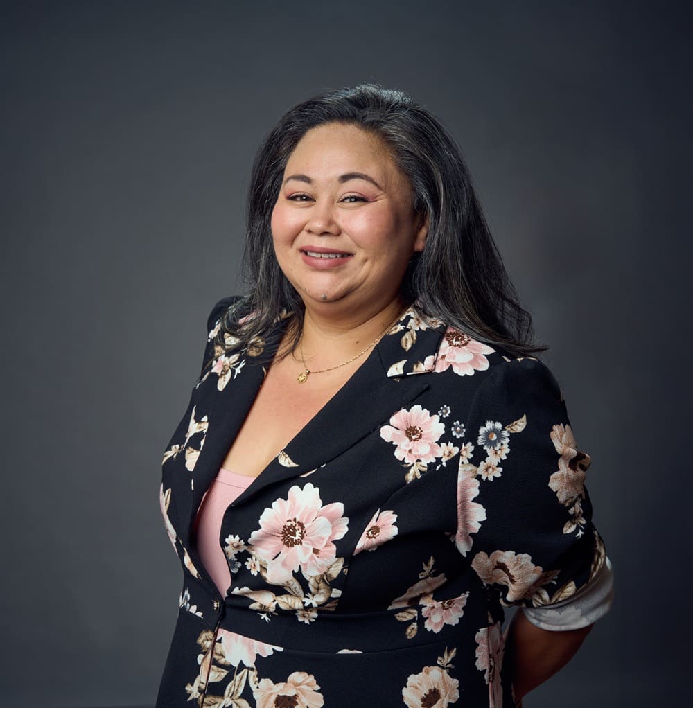 Woman with long dark hair wearing a black floral blazer stands against a plain dark background, smiling with hands behind her back.