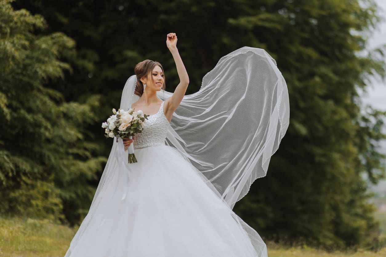 A bride in a white wedding dress holding a bouquet stands outdoors, raising one arm as her veil flows dramatically behind her.
