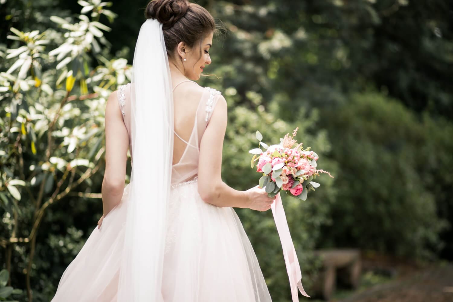 A bride in a white dress with a long veil holds a bouquet of pink flowers while standing outdoors, facing away from the camera.