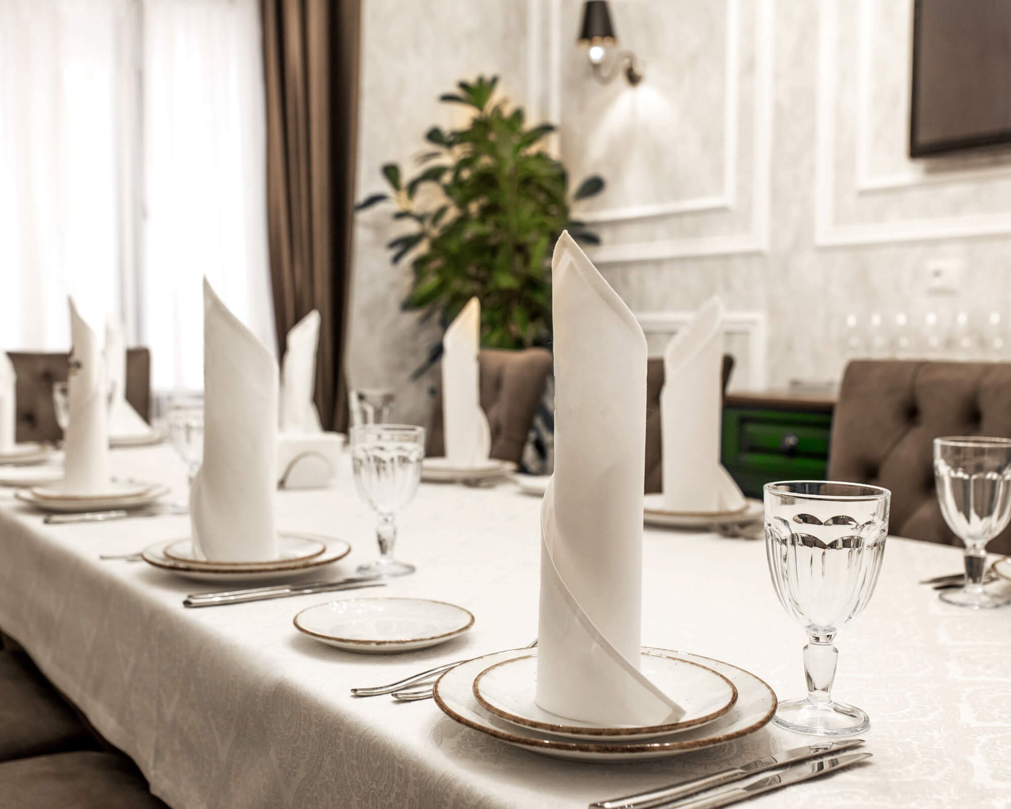 A long dining table set with white tablecloths, folded napkins, plates, and water glasses.